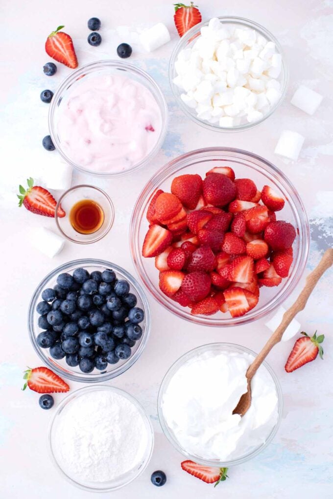 overhead shot of red white and blue cheesecake salad ingredients in bowls on a table featuring fresh blueberries strawberries marshmallows yogurt cream cheese and pudding