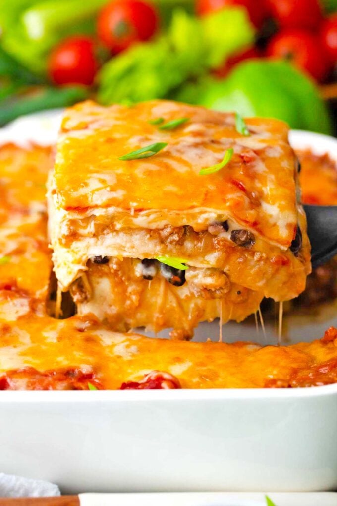 Lifting a slice of cheesy taco lasagna from a casserole dish.