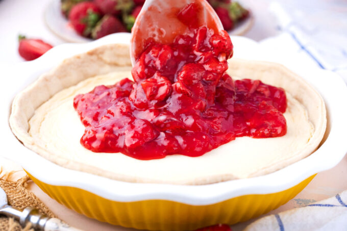 Adding strawberry pie filling on top of cheesecake pie.