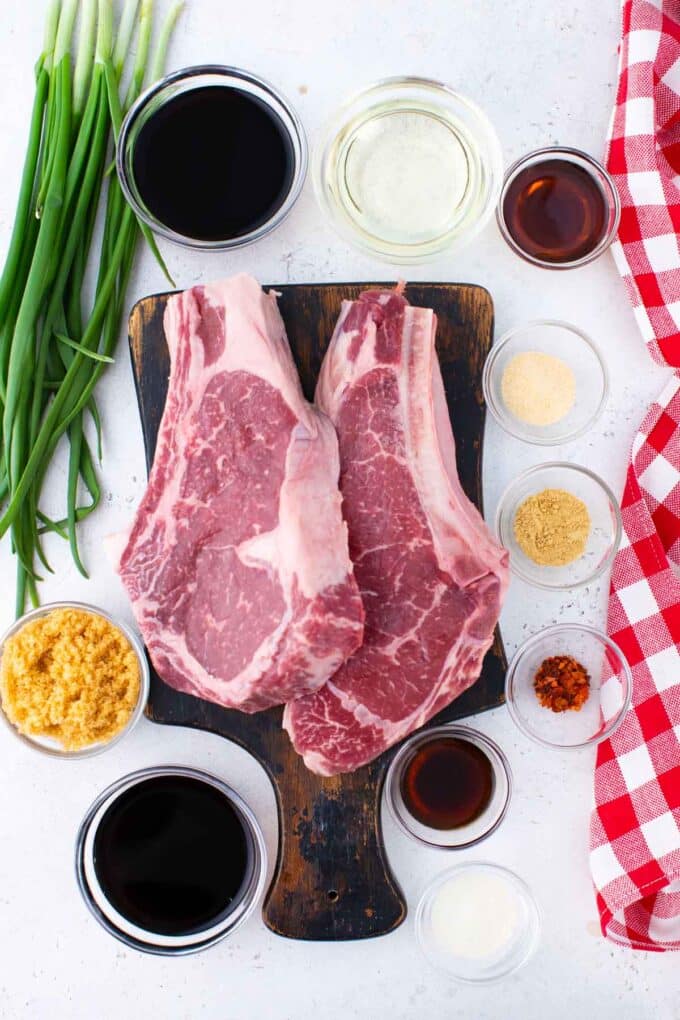 Asian steak marinade ingredients arrange in bowls around a cutting board with two ribeye steaks.