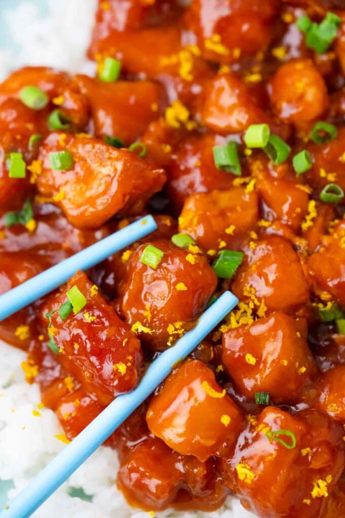 juicy and sticky orange chicken pieces over rice with blue chopsticks next to them