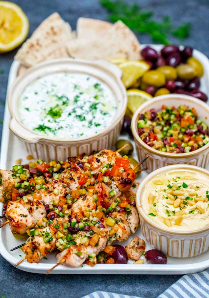 Grilled chicken souvlaki skewers with tzatziki sauce, cucumber, tomato, and olives relish, a bowl of hummus, and pita bread.