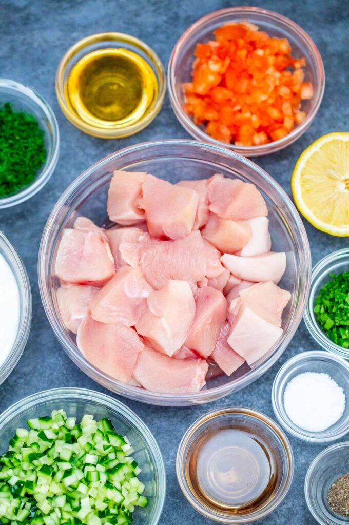 Overhead shot of grilled chicken souvlaki ingredients in bowls on a grey surface.
