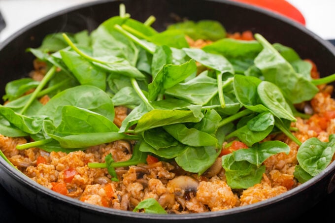 Adding fresh spinach to ground chicken in a skillet.