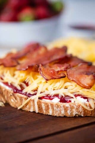 sourdough bread slice topped with cream cheese, strawberry jam, shredded cheddar, and crispy bacon