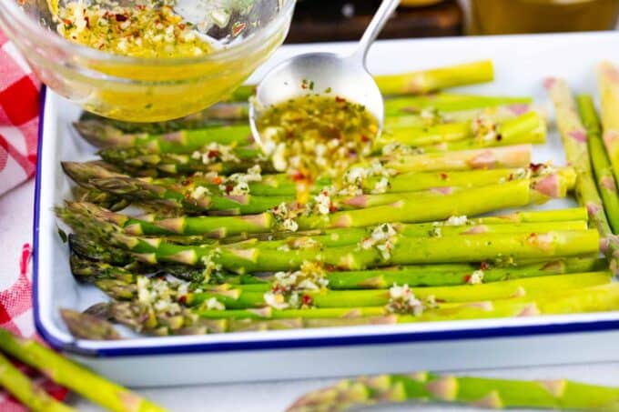 Drizzling olive oil garlic mixture over asparagus spears in a baking pan.