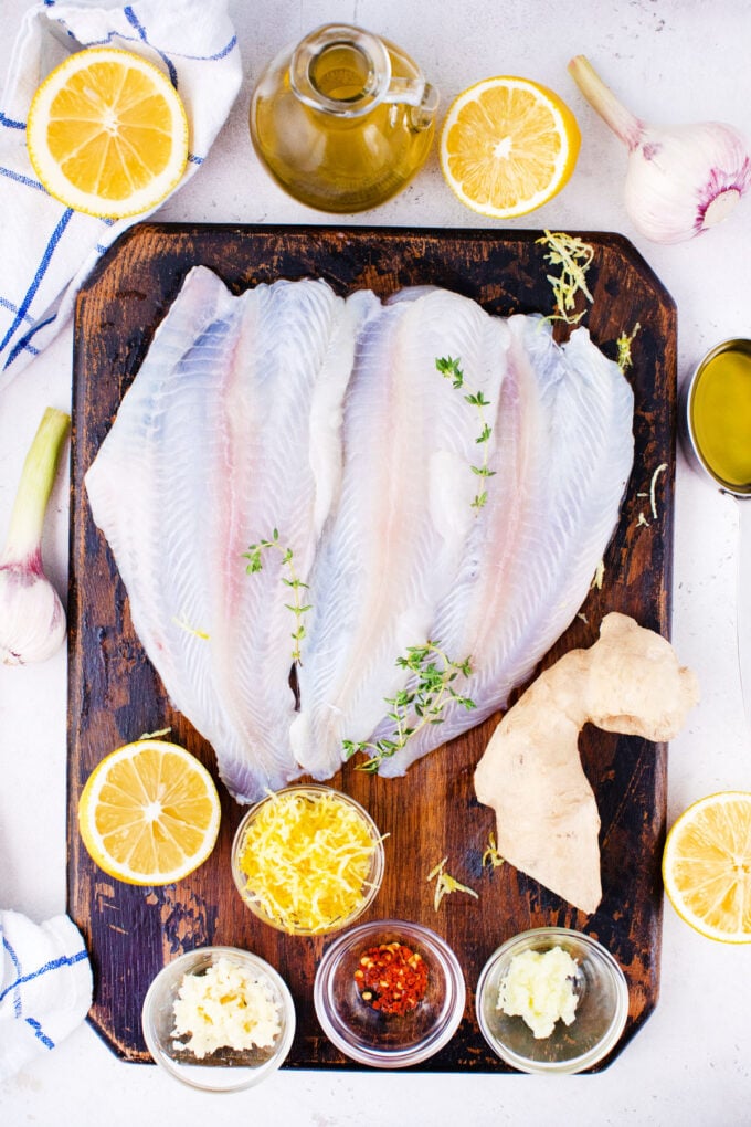 Overhead shot of baked lemon garlic tilapia ingredients in bowls on a white surface.