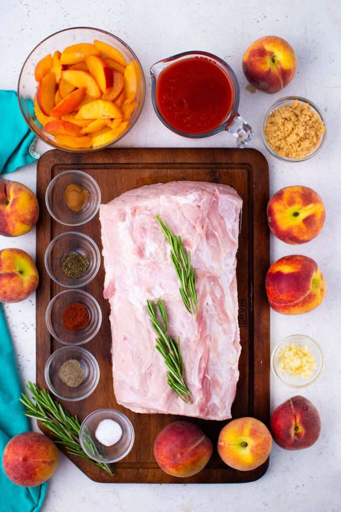 Overhead shot of slow cooker peach pork ingredients in bowls around of a cutting board with a raw pork loin.